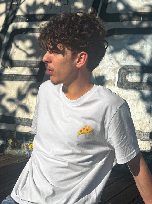 Photo de profil d'un jeune homme aux cheveux bruns bouclés, assis devant un mur recouvert de graffitis. Il porte un t-shirt blanc orné d'un patch brodé en forme de part de pizza.